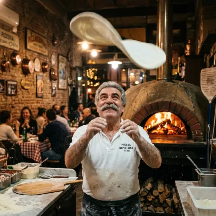 A middle-aged Italian pizzaiolo with a thick grey moustache tossing pizza dough high in the air in a busy Naples pizzeria at night. Motion blur on the dough, glowing wood-fired oven, flour dust, cinematic food photography.