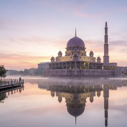 Putra Mosque in Putrajaya at sunrise, its pink dome mirrored perfectly in the calm lake. Pastel sky, soft mist, wide-angle architectural photography.